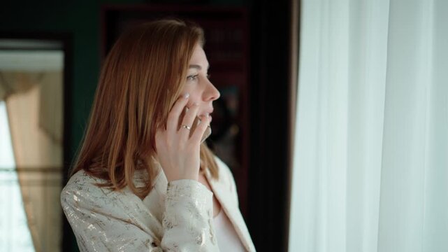 Woman stands by window, gazing outside thoughtfully. She touches face, hand near lips. Her expression shows calm contemplation or mild concern. She wears light-colored blouse