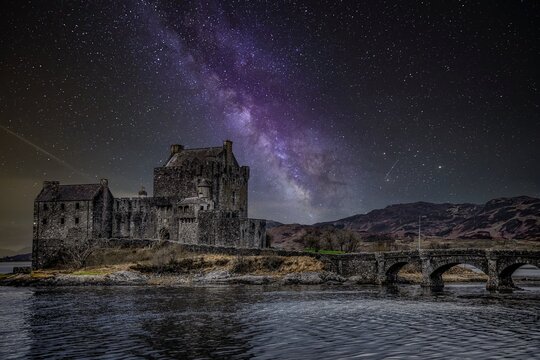 Eilean Donan Castle in Dornie, Scottish Highlands, UK, illuminated at night beneath a starlit Milky Way sky