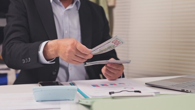 Businessman counts dollar banknotes over financial report papers at office desk. Corporate cash flow management, capital budget investment analysis, commercial banking loan settlement audit.