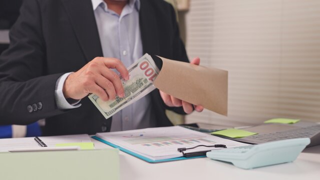 Businessman takes cash dollar bills out envelope behind office desk with financial report. Bribery corruption scandal, corporate kickback payment, under table transaction, illegal loan subsidy.