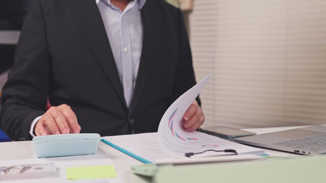 Businessman calculates financial budget data while reviewing charts on paper desk indoors. Corporate audit analysis, tax accounting report, investment profit calculation, commercial paperwork review.
