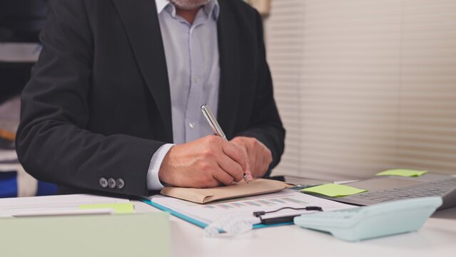 Mature businessman writes on paper envelope over financial report clipboard at office desk. Corporate bribery investigation, corrupt transaction settlement, unofficial bonus payment handling.