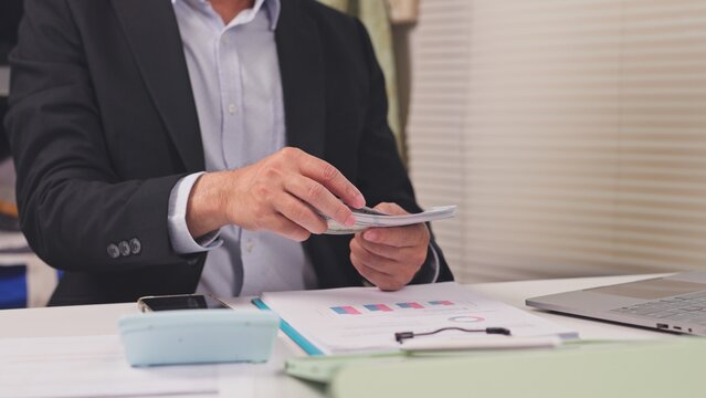Businessman counts bundle cash dollar bills above financial report documents at office desk. Corporate bribery scandal, executive salary payment, investment profit audit, commercial revenue.