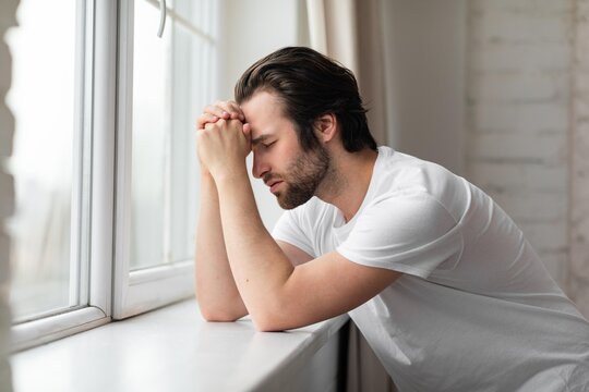 Worried Caucasian Man In His 30s Leaning On Windowsill With Hands Clasped, Deep In Thought At Home, Indoor Setting. Expressing Emotions Reflecting Mental Health And Wellness Concept.