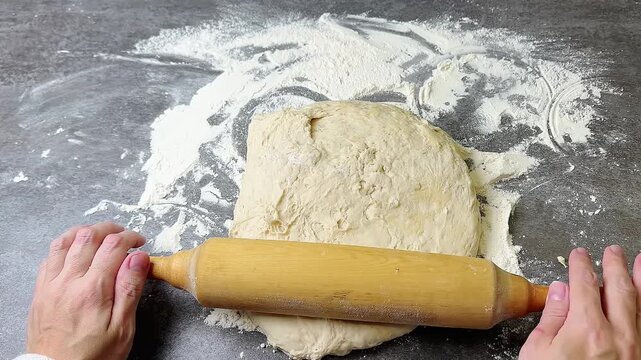 Female hands rolling fresh raw dough with wooden pin on dark stone table dusted with white flour, top view camera zoom in movement, homemade baking preparation process