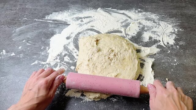 Female hands rolling fresh raw dough with pink wooden pin on dark stone table dusted with white flour, static high angle top view shot, baking preparation process