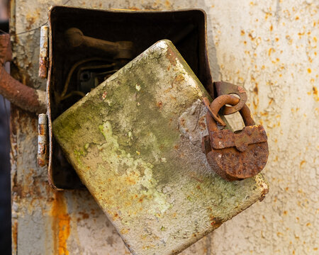 Old rusty padlock hanging on open weathered electrical box door with peeling paint, close up