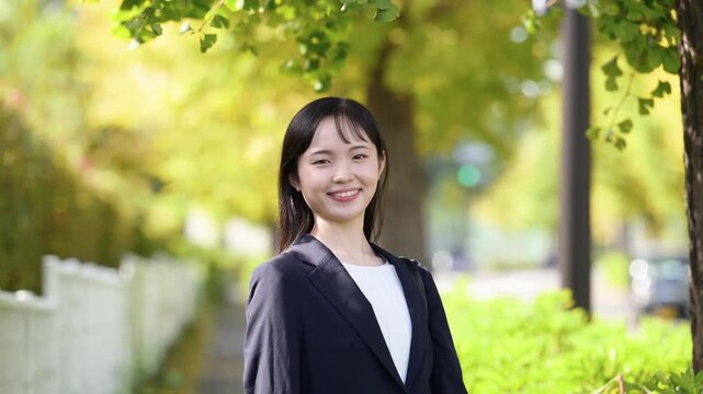秋の紅葉が美しい屋外を笑顔で微笑む、新卒の若い日本人女性社員、新入社員としてフレッシュなイメージ　就職や新生活への期待と希望を胸に前向きに歩く上半身動画カメラ目線で笑顔髪をかき上げる