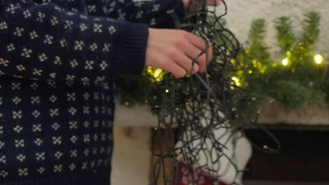 Person in a cozy sweater and plaid pajama pants untangles Christmas string lights near a fireplace with a stocking. Cozy holiday preparation at home