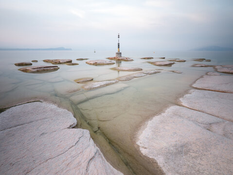 View of the flat, layered rock formations of Jamaica Beach extending into the calm waters of Lake Garda with a navigation beacon in the distance, Sirmione, Lombardia, Italy.