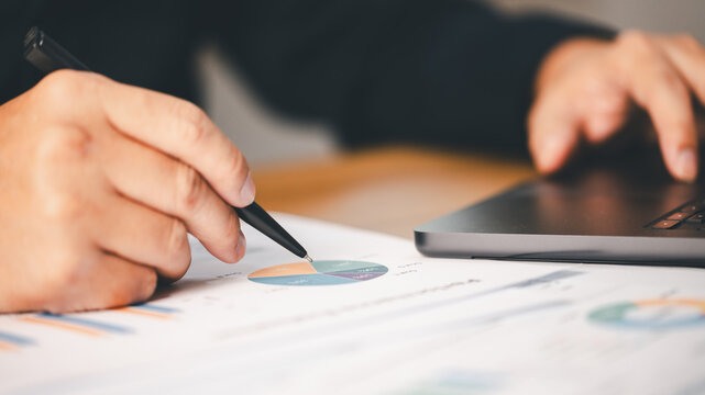 Close-up person holding pen analyzing financial data charts reports working laptop computer desk. Concept business growth strategy corporate investment performance planning office analytics