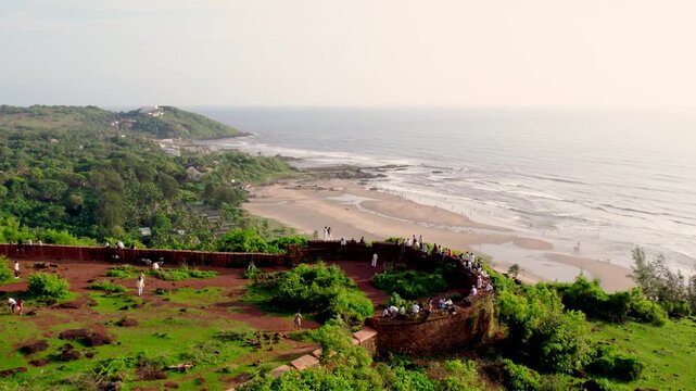 Aerial view of Chapora Fort walls with people sitting on the historic red stone ramparts overlooking the coastline of Vagator Beach in Anjuna, Goa, India.