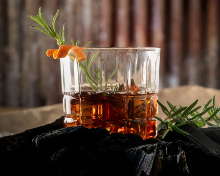 Old fashioned cocktail in a whisky glass with a sprig of fresh rosemary and orange peel, sitting on a bed of chunk charcoal