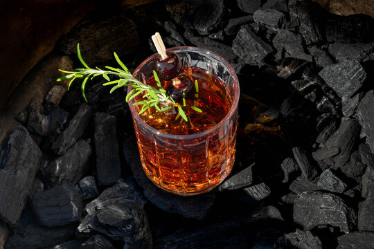 Old fashioned cocktail in a whisky glass with a sprig of fresh rosemary and cherries, sitting on a bed of chunk charcoal