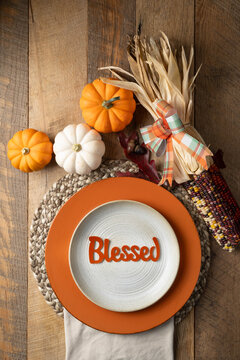 Rustic table setting with colorful pumpkins and corn accents with the word, Blessed on a stoneware plate with orange riser and woven placemat.