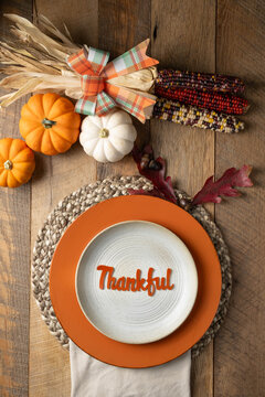 Rustic table setting with colorful pumpkins and corn accents with the word, Thankful on a stoneware plate orange riser and woven placemat.