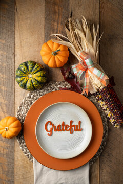 Rustic table setting with colorful pumpkins and corn accents with the word, Grateful on a stoneware plate with orange riser and woven placemat.