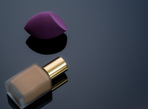 Purple makeup sponge with beige liquid foundation bottle on a dark reflective surface, studio beauty product still life for cosmetic application, makeup routine, skincare branding and advertising.