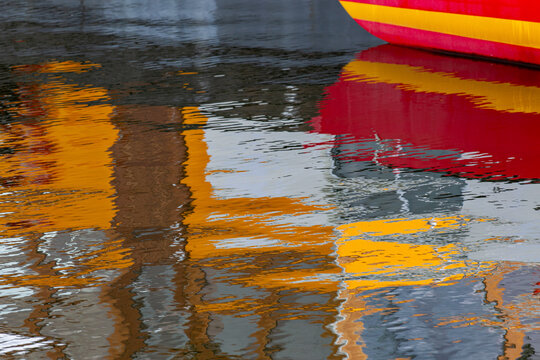 View of the colorful reflection of the Sluishuis building and a red and yellow boat in the rippling water of the IJ canal in Amsterdam, North Holland, Netherlands.