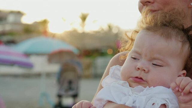 Close-up of a curly-haired baby girl with blue eyes in her mother's arms on a beach at sunset, with parasols blurred behind, French Riviera, France.