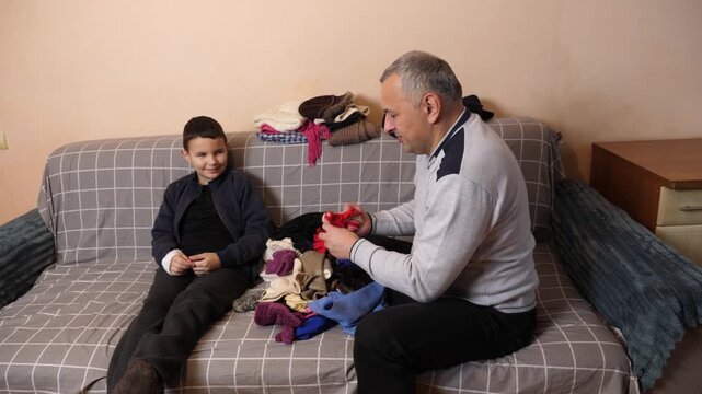 A father sorts warm clothes with his son, showing him red knitted socks and examining garments among a pile of winter clothing.