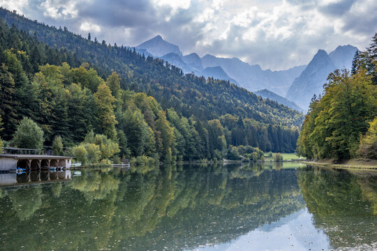 View of Riessersee lake surrounded by dense autumn forests and towering Alpine mountains under a cloudy sky in Garmisch-Partenkirchen, Bavaria, Germany.