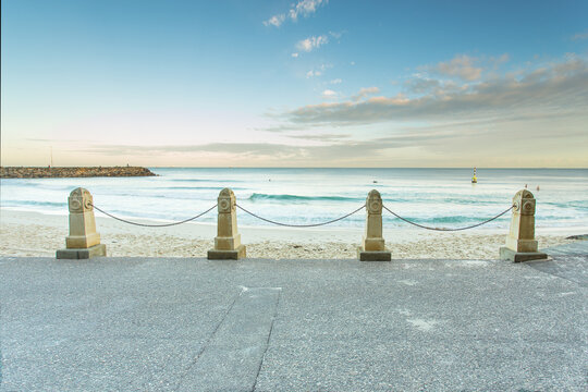 View of Cottesloe Beach with stone bollards and chains along the promenade overlooking the calm ocean under a clear sky in Cottesloe, Western Australia, Australia.