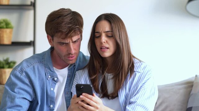 Confused young couple looking at a smartphone screen, having a serious disagreement and feeling frustrated while sitting together on the sofa in their modern living room apartment