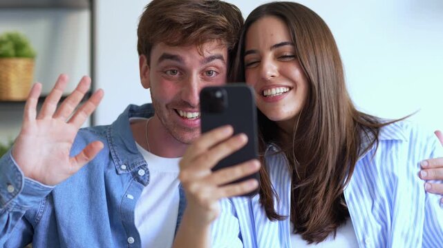 Cheerful couple sitting at home making a video call on a smartphone, waving hello and smiling. The affectionate partners are using their mobile device for online communication with family