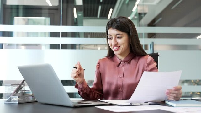 Happy businesswoman sitting in office, looking at laptop screen with satisfaction after checking financial results. Smiling financier pleased with profit and growth, celebrating success in workplace.
