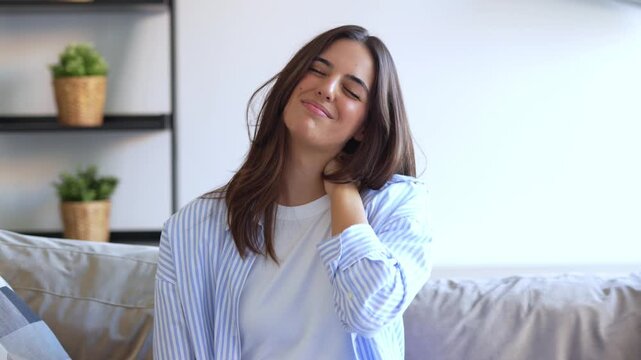 Exhausted beautiful woman massaging her stiff neck and tense shoulders after a long day of work while sitting on the sofa in her living room, trying to relieve the muscle pain and fatigue