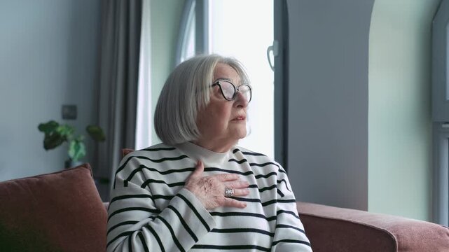 Worried senior woman with grey hair experiencing a sudden health problem, feeling chest pain, and shortness of breath while sitting on the sofa in her living room, a possible heart attack