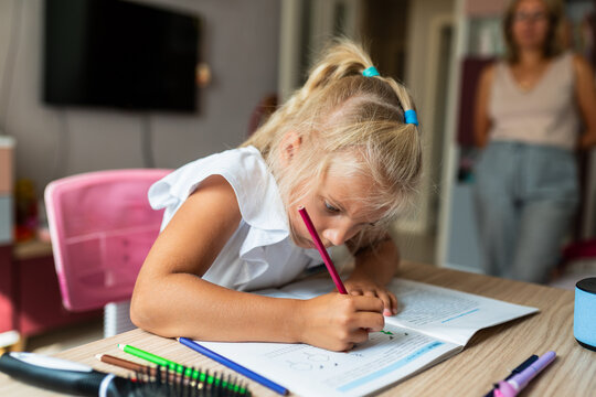 Young schoolgirl completes homework independently while her mother watches. Child learning at home, parental supervision and school education