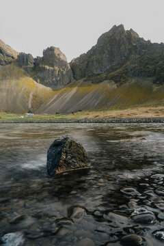 View of a dark rock in a flowing stream with rugged mountains and mossy slopes in the background in Sveitarfelagid Hornaf, Iceland.