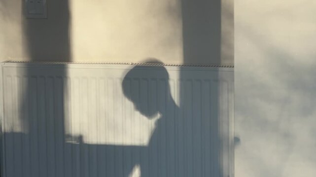 A boy's bowed head appears as a mealtime shadow on a sunlit wall above a radiator.