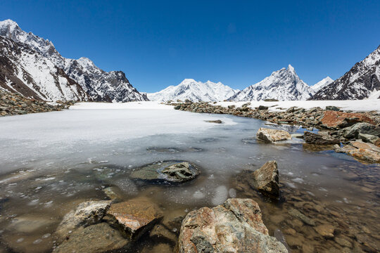 View of the majestic K2 mountain surrounded by snow-capped peaks, with a partially frozen alpine lake and rocky shoreline in the foreground, Pakistan.