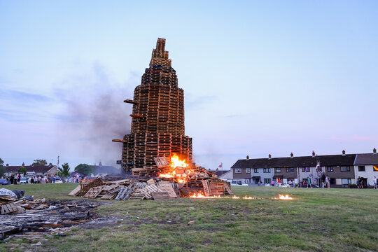 View of a large bonfire constructed from stacked wooden pallets burning in a residential field with houses in the background in Larne, Northern Ireland, United Kingdom.