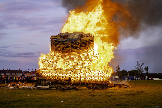 View of a large bonfire made of wooden pallets burning brightly at dusk, with a crowd of people gathered in the background on a grassy field in Larne, Northern Ireland, United Kingdom.