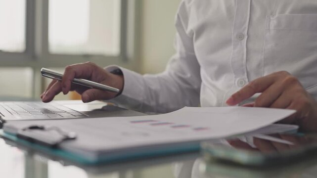 Businessman reviews financial chart paperwork while typing on laptop keyboard in office. Corporate audit analysis, market research data evaluation, quarterly revenue investment report management.