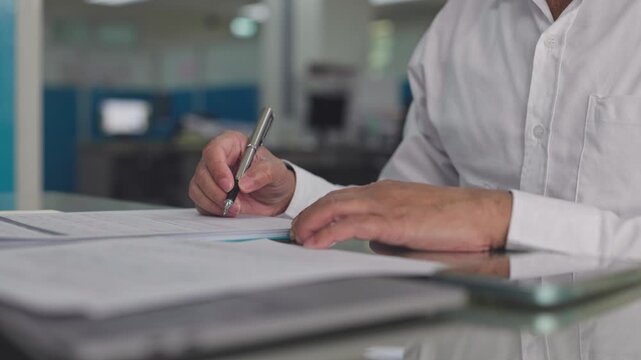 Businessman writes notes on paper form beside tablet computer in workplace. Corporate contract signature, financial audit review, legal agreement paperwork analysis.