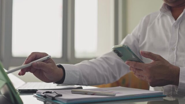 Businessman points stylus toward tablet screen while holding mobile phone beside paperwork on desk. Corporate financial audit analysis, multitasking workflow management, digital investment review.