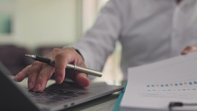 Businessman reviews financial audit report document beside laptop computer in office. Corporate accounting inspection, annual investment analysis, market performance evaluation data review.