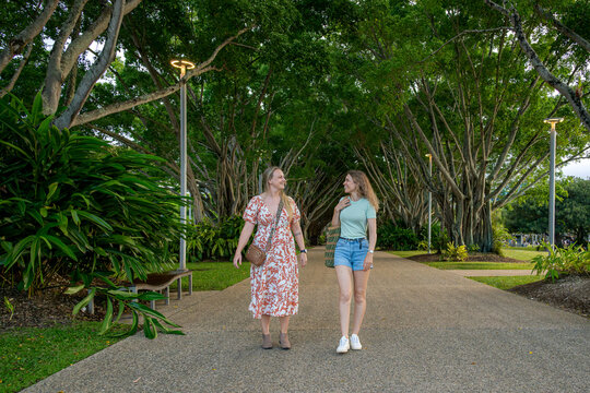 Two women walking side by side along a tree-lined pathway in a lush park