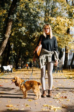 A woman with glasses walks a leashed brown dog along a paved path in a park. Fallen leaves cover the ground, and other people and trees are visible in the background