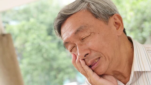 60s senior east asian man suffering toothache indoors, holding his cheek with a painful grimace in natural light