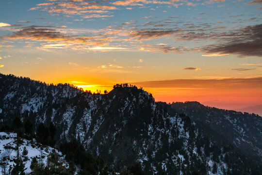 View of Malam Jabba mountain range covered in snow during a vibrant sunset with golden light illuminating the clouds above the peaks in Sawat, Khyber Pakhtunkhwa, Pakistan.