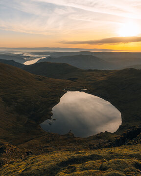 View of Red Tarn nestled below the slopes of Helvellyn at sunset with rolling hills and a golden sky in Keswick, England, United Kingdom.