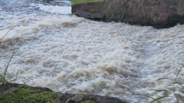 A close view shows white foam swirling against a stone wall downstream from a small city weir. 