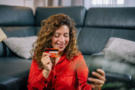 Smiling woman holding credit card near mouth looking at smartphone