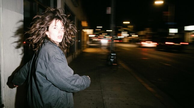 Young woman with windblown hair looking back on a city sidewalk at night beside blurred traffic and illuminated buildings in an urban street scene